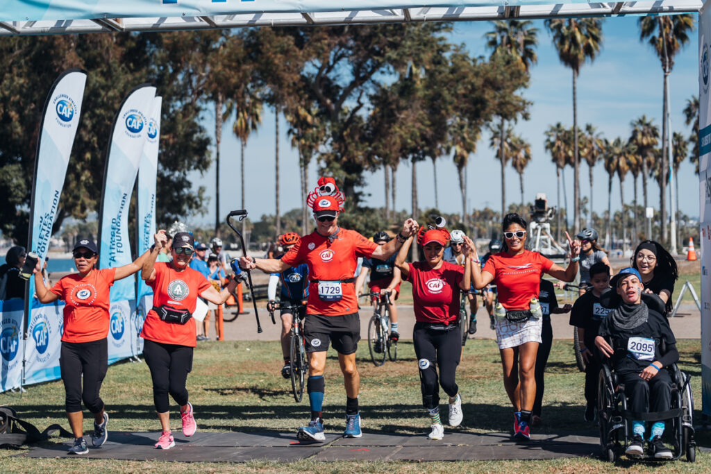 Participants in red shirts cross the finish line arm-in-arm at a vibrant, inclusive outdoor event.