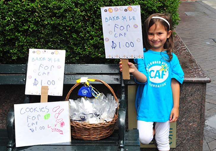 Young girl in a CAF shirt holding a handmade sign for a $1 bake sale fundraiser with cookies displayed in a basket