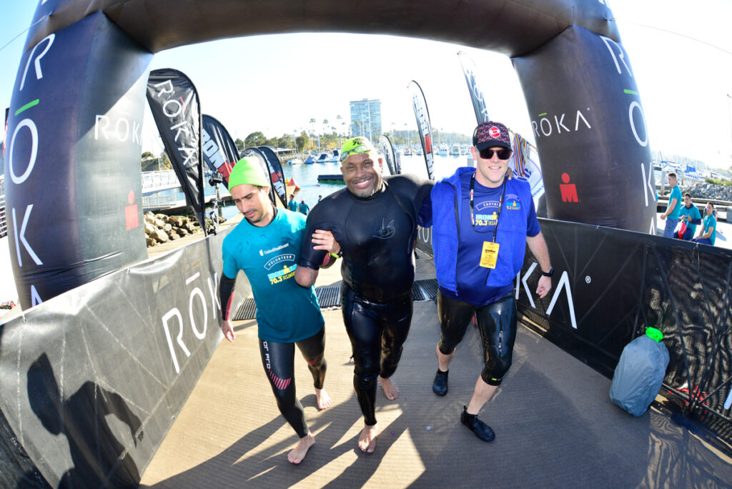 Three people walking under a ROKA archway near a marina, with one smiling widely.