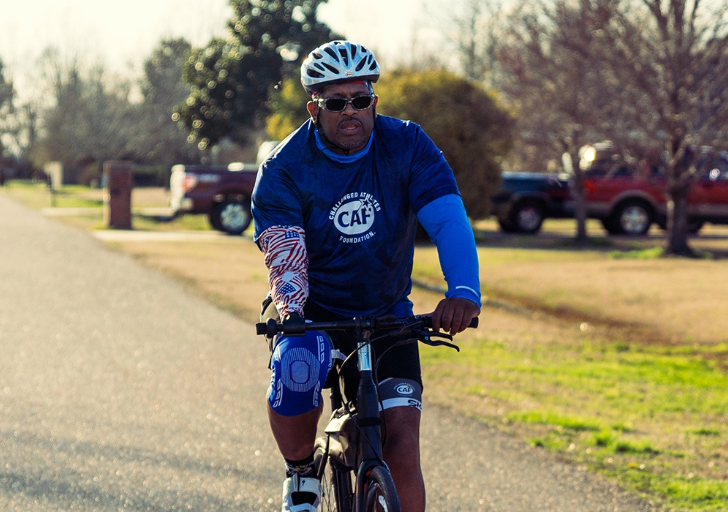 A person with an upper limb difference cycling on a suburban street, wearing a blue shirt, white helmet, and sunglasses.