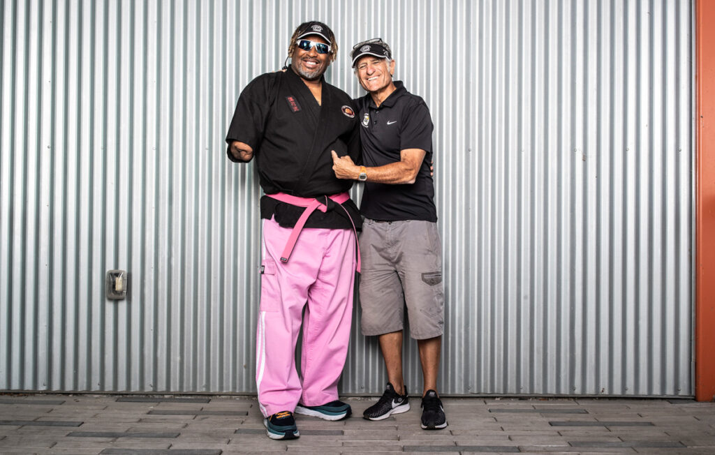 Two people posing together in front of a corrugated metal wall, one in martial arts attire and the other in casual wear.