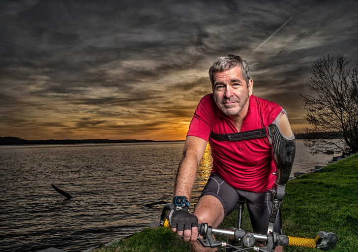 Miles O’Brien, a triathlete with a prosthetic arm, poses on his bike by the water at sunset.