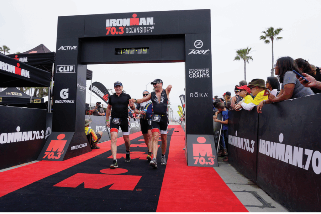 Miles O’Brien crosses the Ironman 70.3 Oceanside finish line holding hands with teammates as spectators cheer.