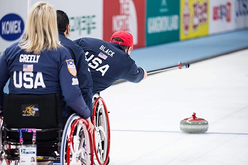 Two Team USA wheelchair curlers competing, one releasing a stone with a delivery stick.