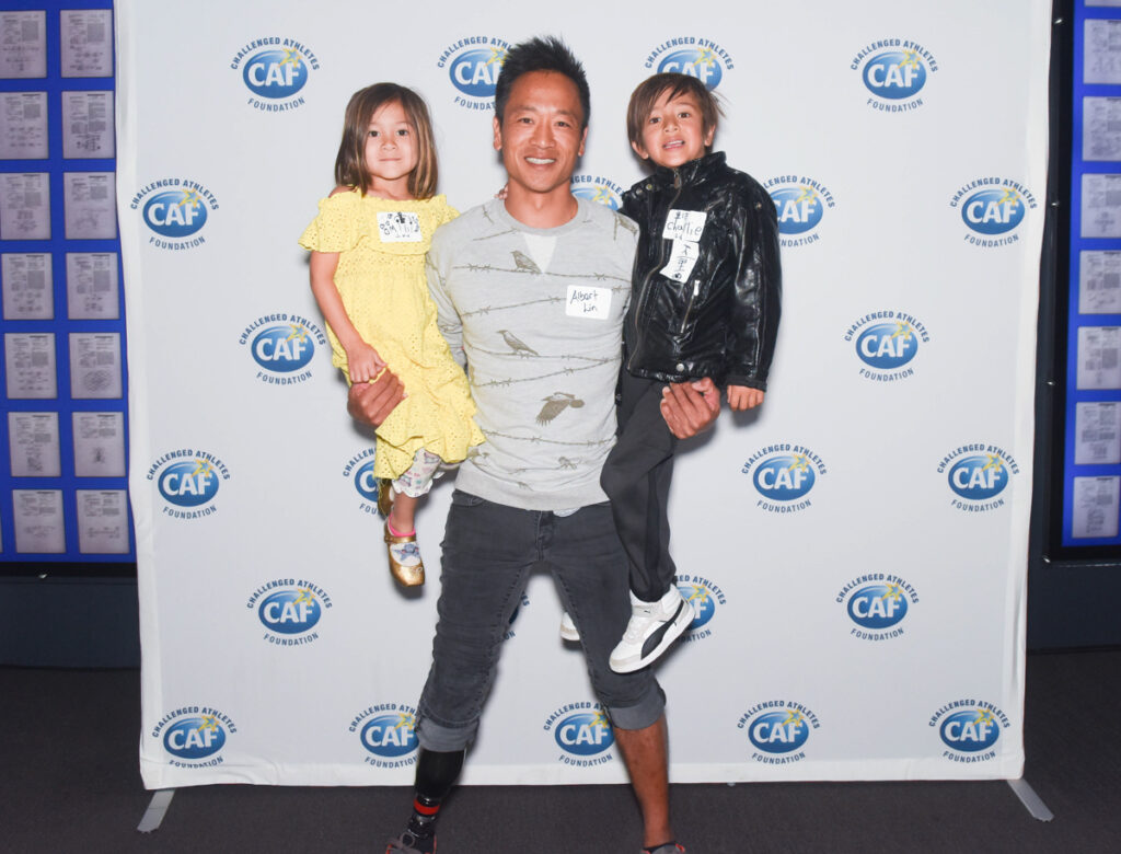 A man with a prosthetic leg holding a young child in a yellow dress and another child in a black jacket, against a Challenged Athletes Foundation backdrop.