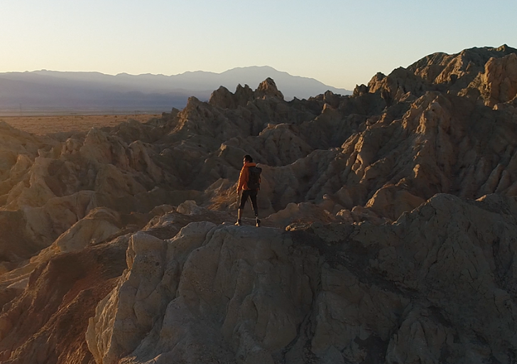 A person in an orange jacket stands on jagged rocks with a distant mountainous background.