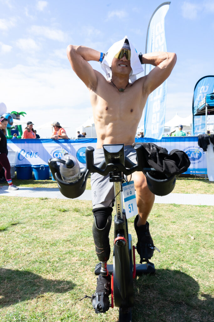 Person with a prosthetic leg exercising on a bike, using a towel to cover their head, surrounded by event banners and people.
