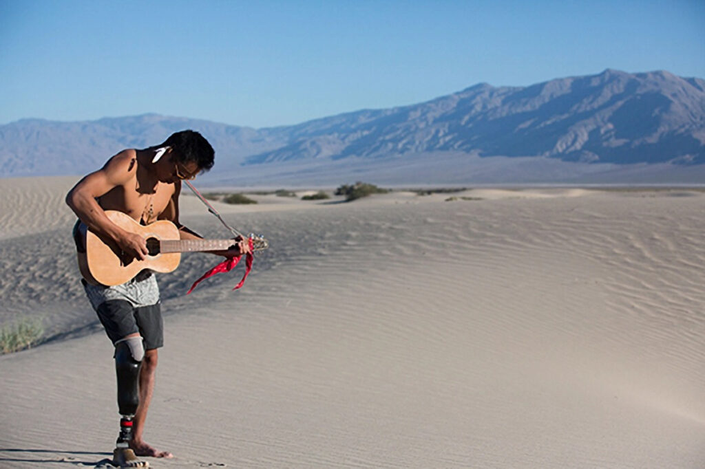 A person with a prosthetic leg playing an acoustic guitar amidst desert dunes with mountains in the background.