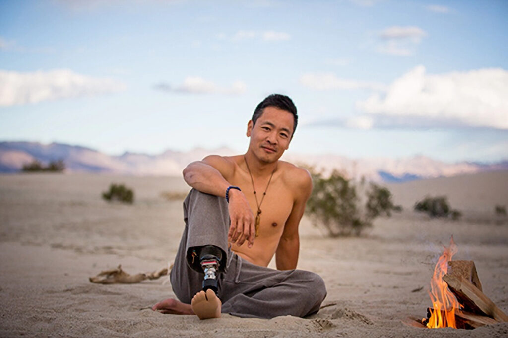 A shirtless man with a lower limb difference sitting on sand near a campfire in a desert landscape with mountains in the background.