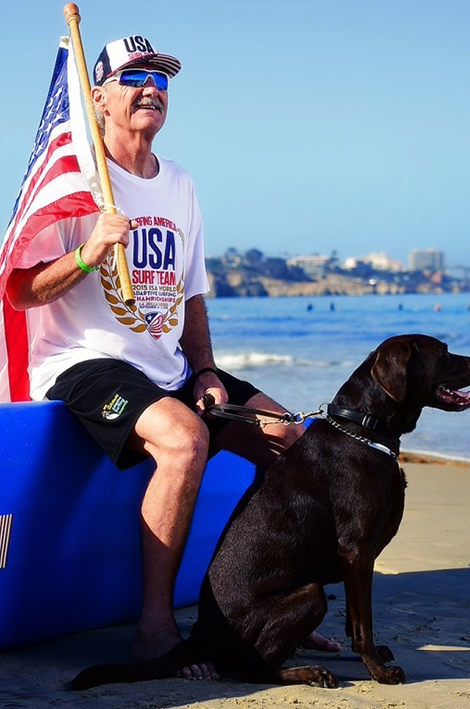 A blind athlete wearing a USA Surf Team shirt sits on the sand holding an American flag with a guide dog beside them, looking toward the water.