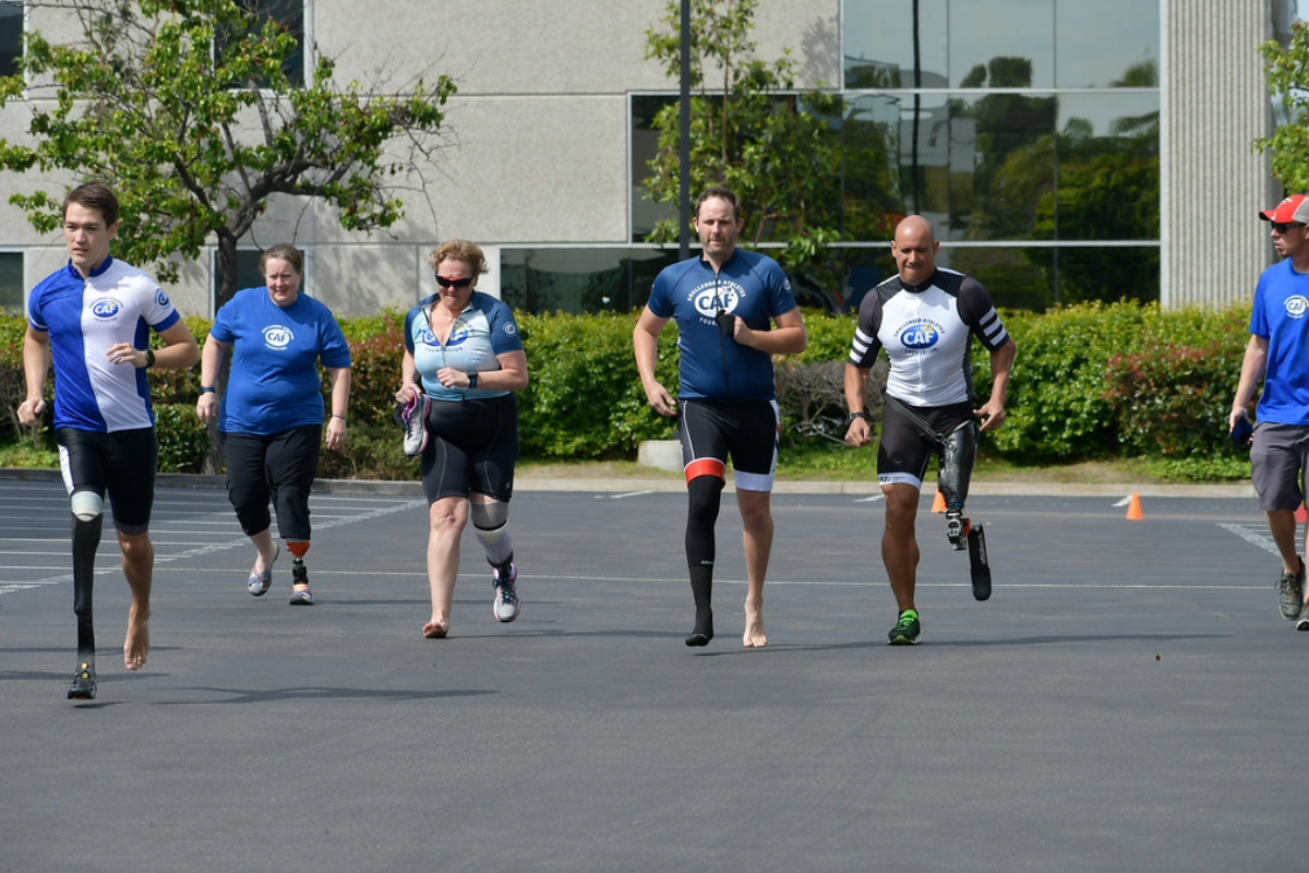 Group of paratriathletes with prosthetic legs and supporters running together in a parking lot during training.