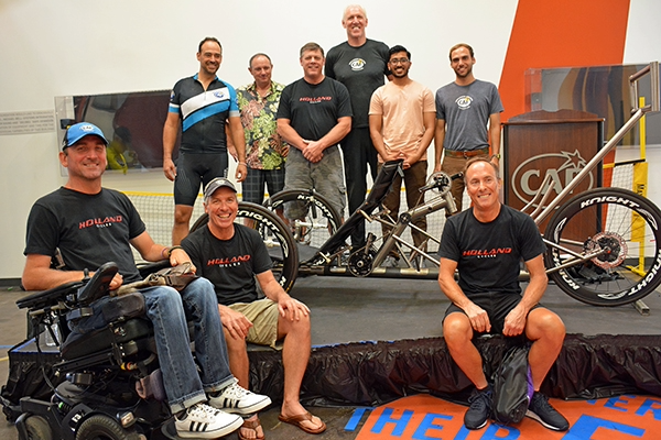 Group of athletes and engineers gathered around a custom tandem handcycle inside a training facility.