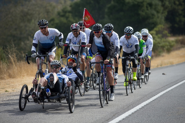 Team of cyclists riding in formation alongside a handcyclist on a rural road during a long-distance event.