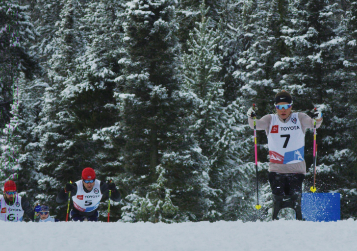 Athletes compete in a cross-country ski race through a snowy forest, with one sit-ski athlete, Oksana Masters, leading the group.