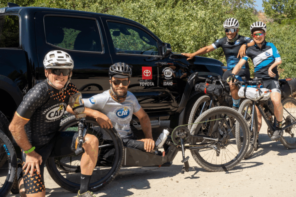Four cyclists, including a handcyclist, pose in front of a black Toyota truck with CAF and Idaho logos during Smoke-n-Fire 400.