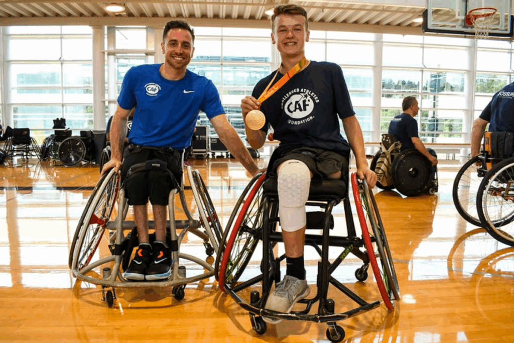 Steve Serio and a young athlete both in sport wheelchairs smile on an indoor basketball court, the young athlete is proudly holding up Steve's gold medal