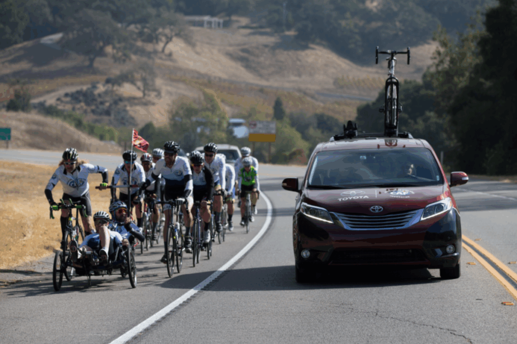 A large group of cyclists, including handcyclists, rides together on a highway next to a Toyota support vehicle during the Million Dollar Challenge.