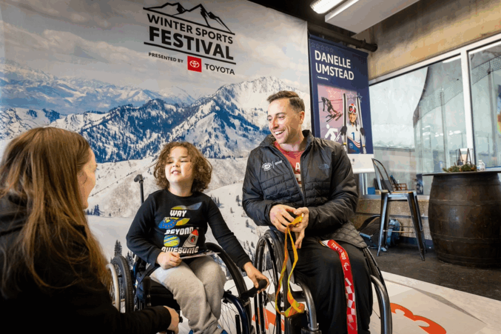 Steve Serio and a young boy in a wheelchair smile and talk with a woman at the Winter Sports Festival, presented by Toyota, with snowy mountain scenery in the background.