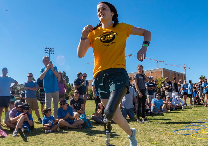 Athlete with a running blade sprints across a grass field during an adaptive sports clinic, with a crowd watching in the background.