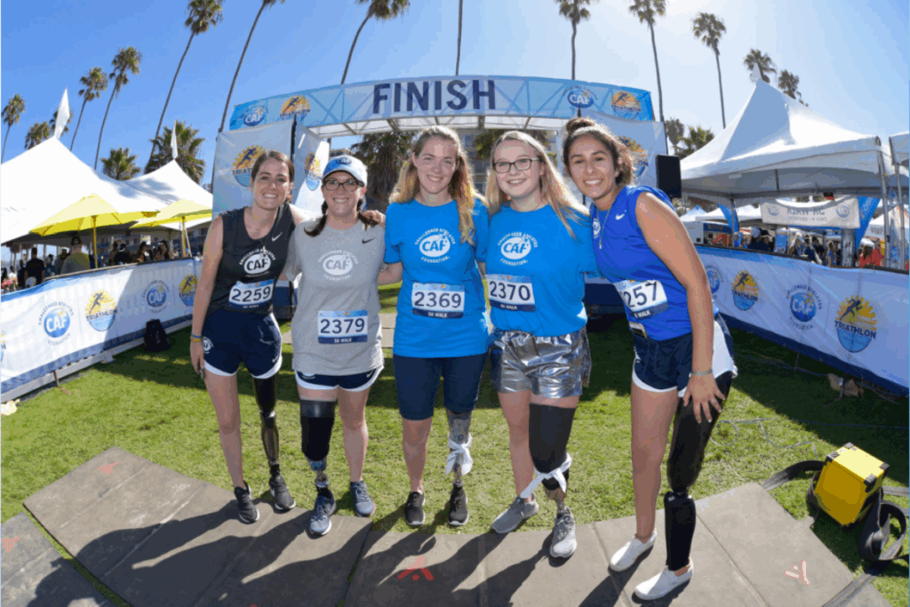 Group of young athletes, some with prosthetic legs, stand arm in arm on a low wall at the beach, smiling toward the camera