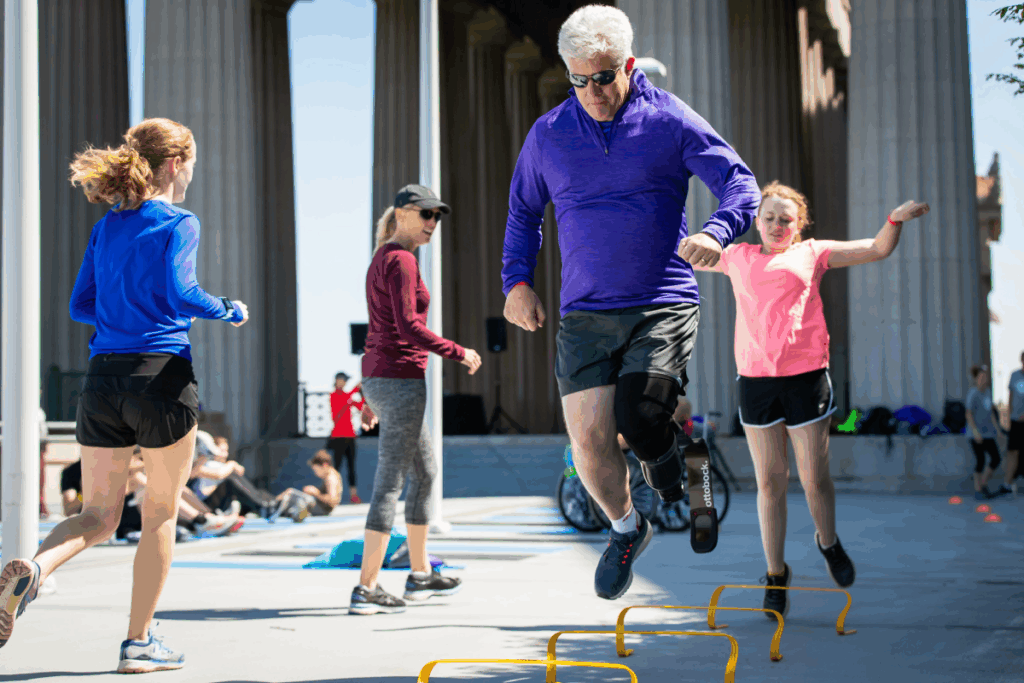 A participant with a prosthetic leg jumps over small hurdles during an outdoor fitness session, with others following behind.
