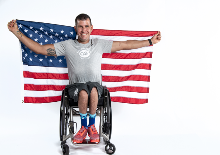 A wheelchair athlete smiles while holding a large U.S. flag behind him in a studio portrait.