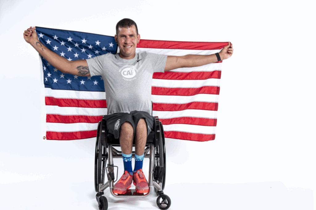 A wheelchair athlete smiles while holding a large U.S. flag behind him in a studio portrait.