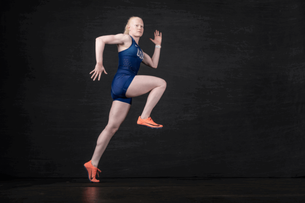 A track sprinter in a USA uniform poses mid-stride against a dark studio background, showcasing powerful running form.