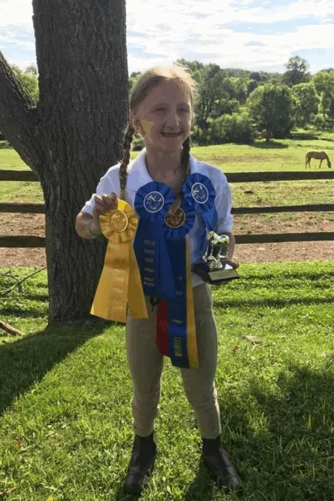 A young equestrian smiles while holding multiple blue and yellow first-place ribbons and a small trophy.