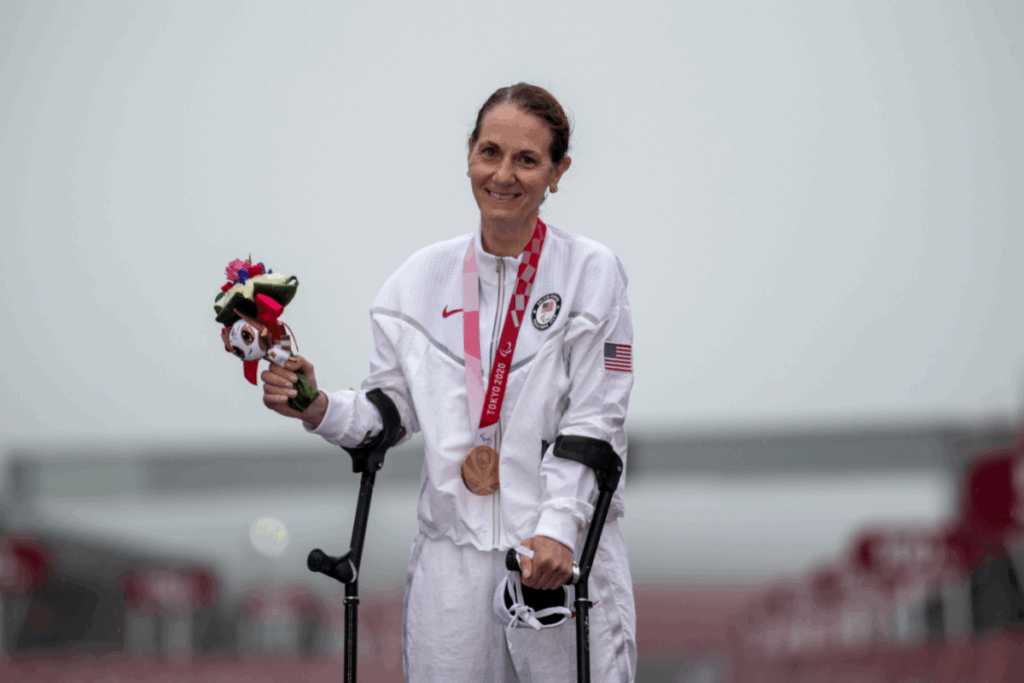 Paralympic athlete wearing Team USA gear smiles with a bronze medal and mascot while standing with crutches on the podium.