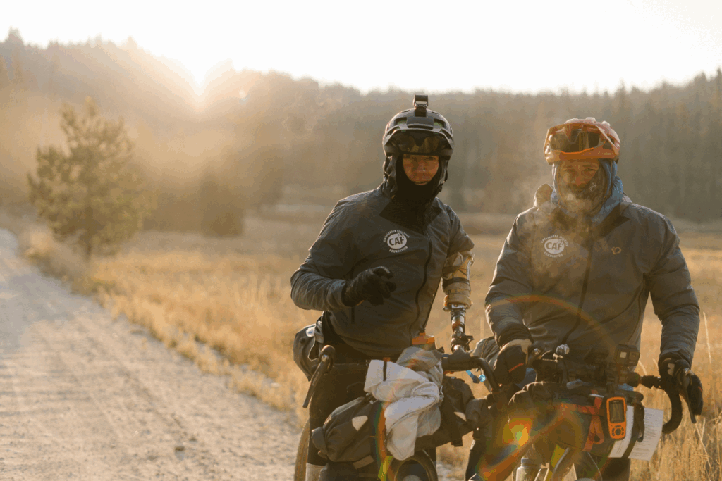 Two adaptive cyclists wearing cold-weather gear and helmets stand beside their loaded bikes at sunrise on a dirt road.