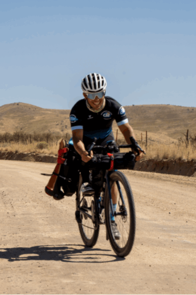 An adaptive cyclist rides a gravel bike on a dirt road, smiling under a clear blue sky.
