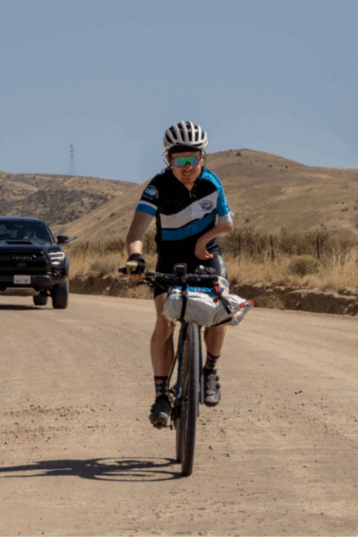 An adaptive cyclist rides toward the camera on a dirt road with a support vehicle behind them.