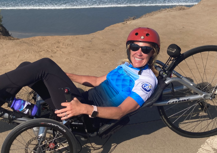 A woman wearing a red helmet and CAF cycling jersey smiles while resting on her handcycle near the beach.