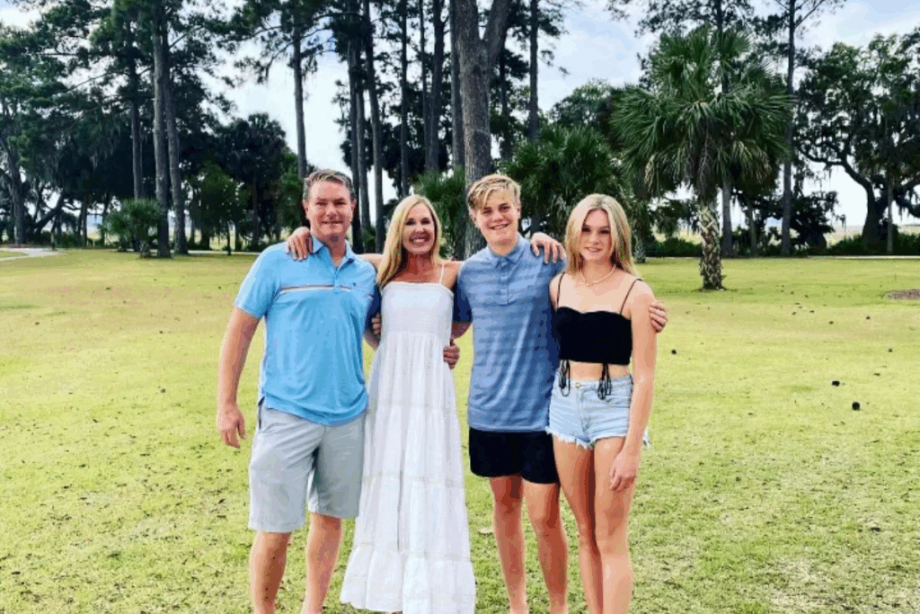 A family of four stands together on a grassy field with trees in the background, smiling for a group photo.