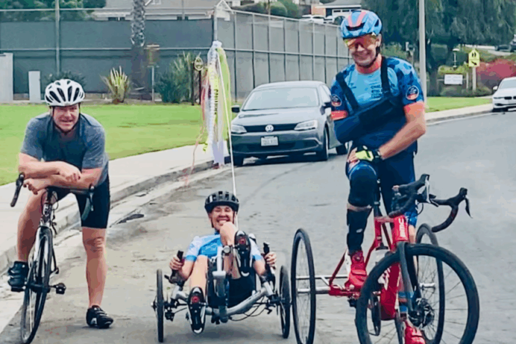 Three cyclists, including a handcyclist in the center, pause and smile on a residential street during a ride.