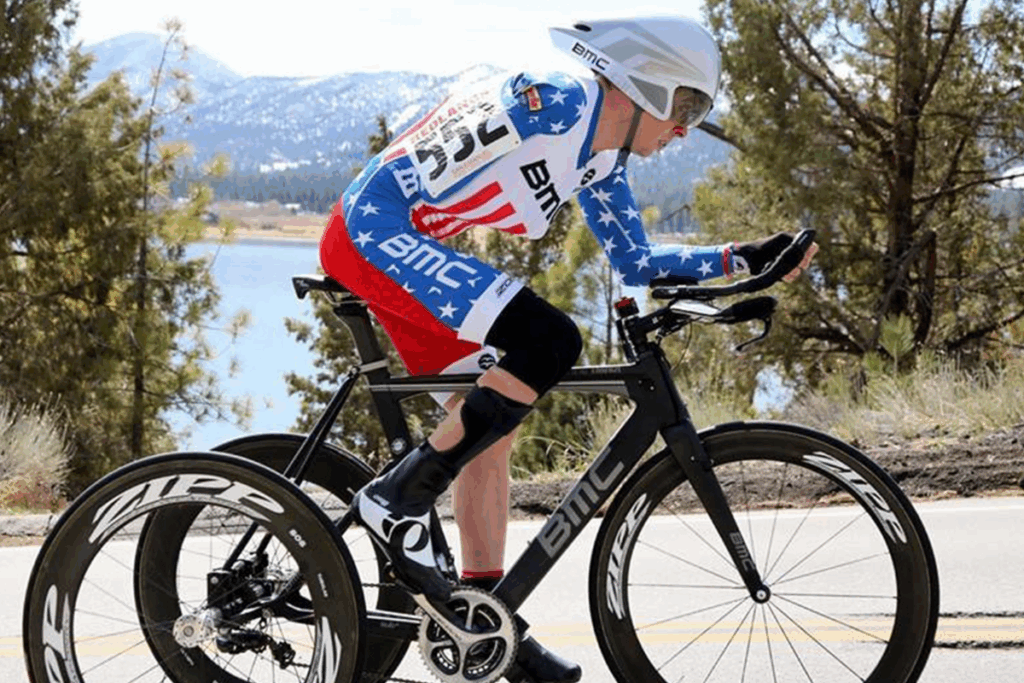 A cyclist wearing a stars and stripes skinsuit and aerodynamic helmet rides a time trial bike on a scenic road near a lake and mountains