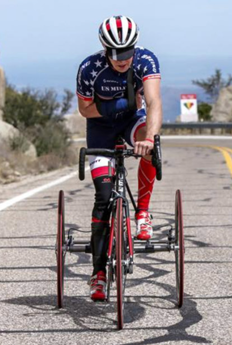 Paralympic cyclist wearing a US Military jersey rides a red adaptive trike uphill on a mountain road.