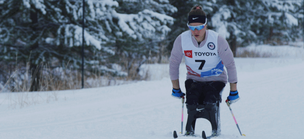 A sit-skiing athlete wearing a number 7 bib on a snowy trail with trees in the background.