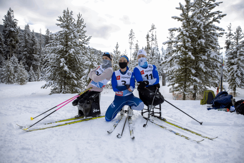 Three individuals in adaptive skiing equipment pose in a snowy forest with ski gear, surrounded by snow-covered trees.