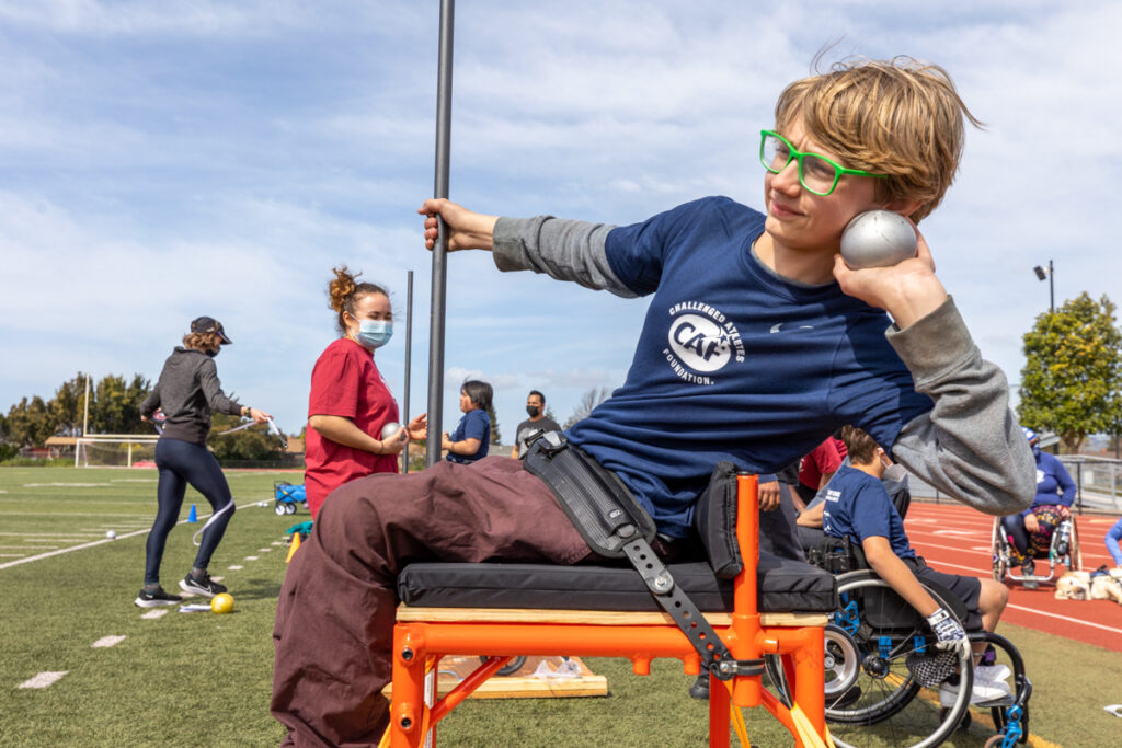 A young person in green glasses prepares to throw a shot put from an adaptive chair on a sports field.