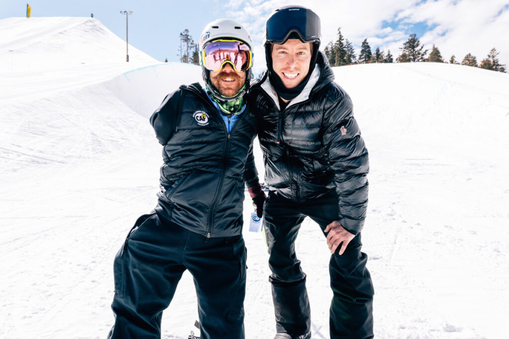 Two people in winter gear smiling on a snowy slope with a clear sky. One of them is Shaun White. The other has a visible upper limb difference.