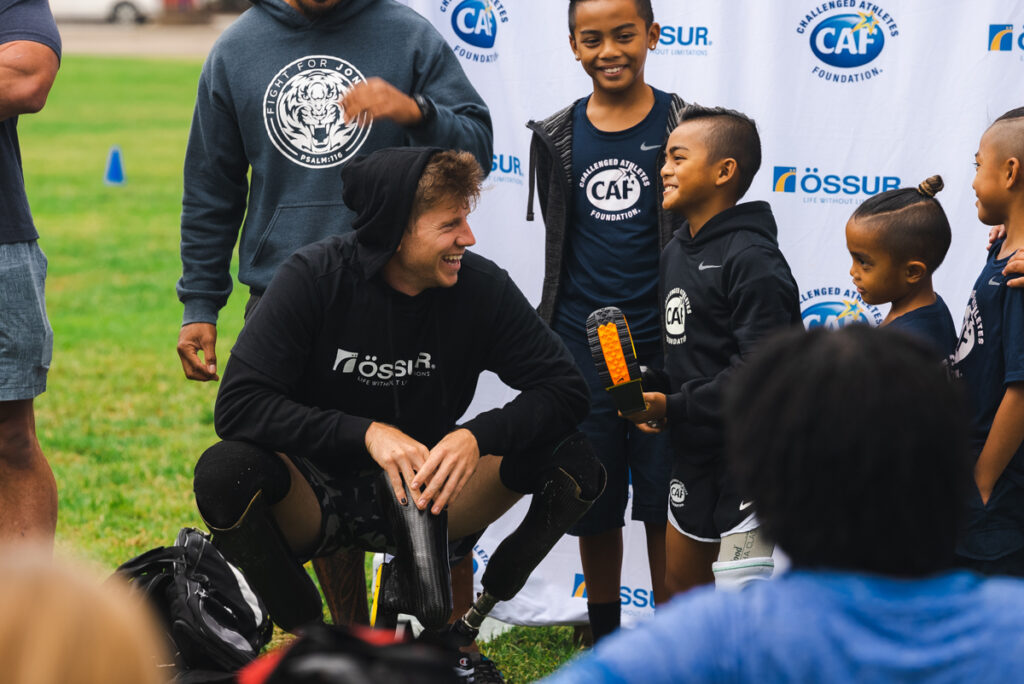 A man with prosthetic legs kneels on grass, smiling at children holding a trophy, under a backdrop with "CAF" and "Össur" logos.