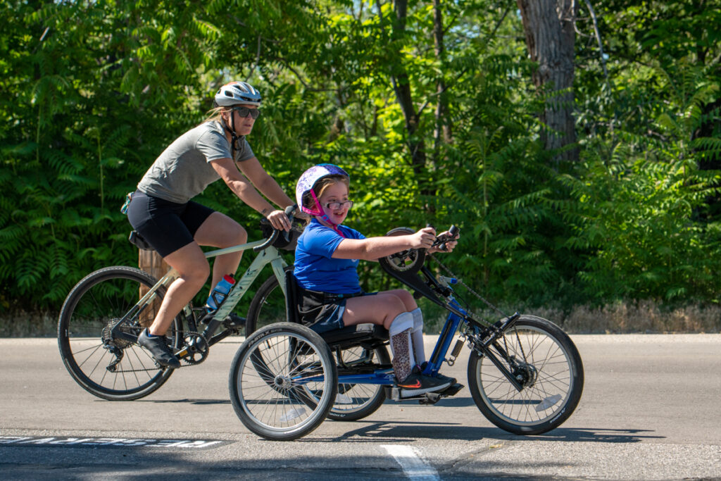 A child on a recumbent tricycle and an adult on a bicycle ride together on a sunny day.