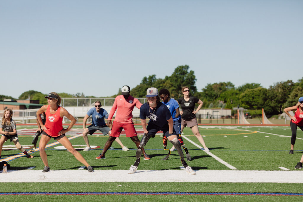A group of diverse people, including individuals with prosthetic limbs, performing warm-up exercises on a green field under a clear blue sky.