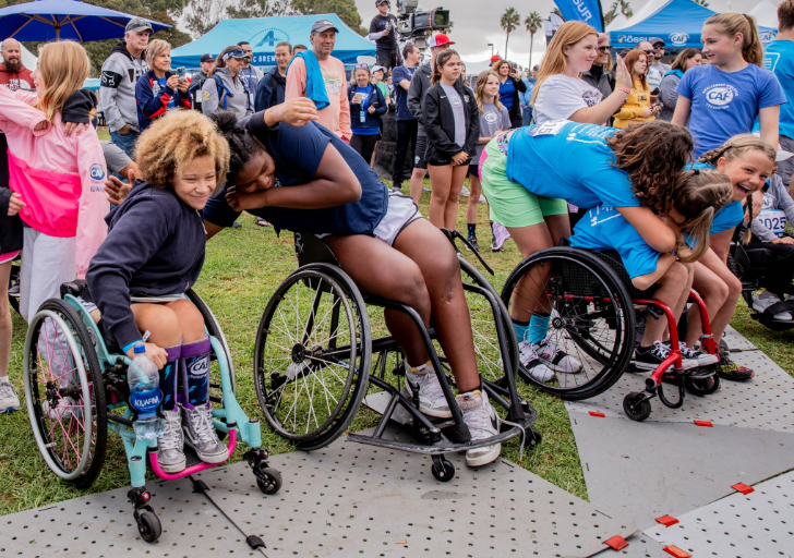Four young athletes in wheelchairs hug each other at a sporting event, surrounded by onlookers and tents on a grassy area.