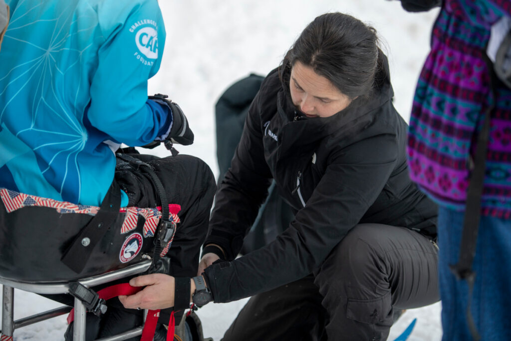 A person assists another in a specialized chair on snow.