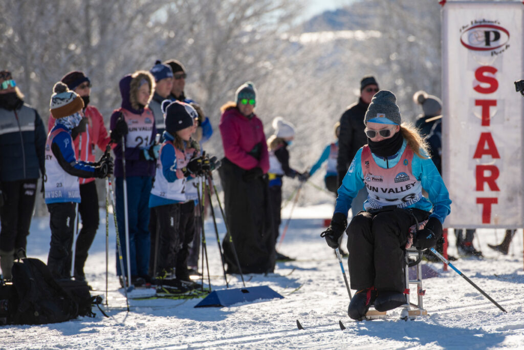 Participants at a snowy race start, including a person using adaptive skiing equipment near the "START" banner.