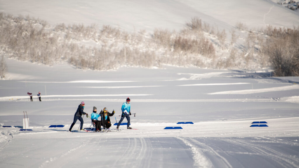 A group assists a sit-skier on a snowy cross-country trail with snow-covered trees in the background.