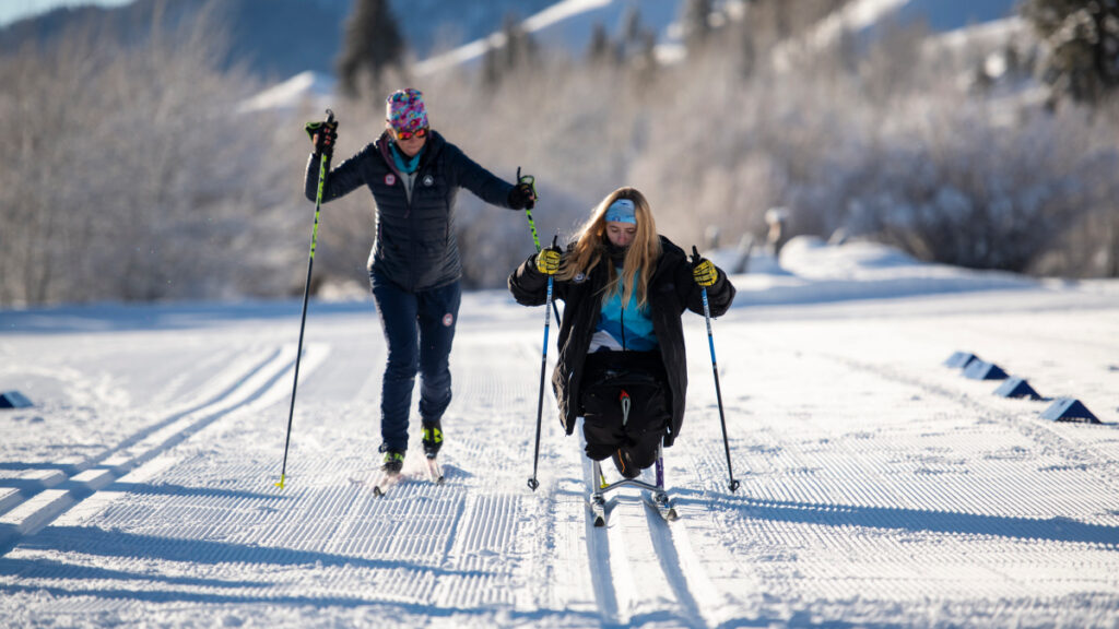 Two people skiing on a snowy trail, one using a sit-ski.